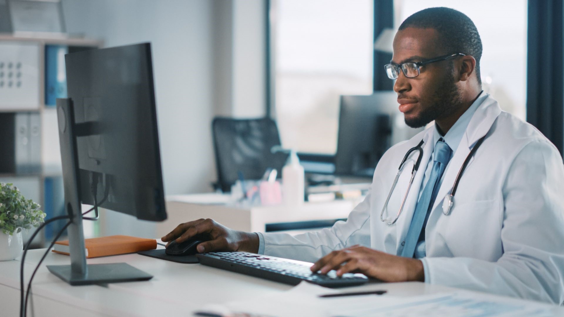 A doctor looks seriously at a computer screen while holding keyboard and mouse.