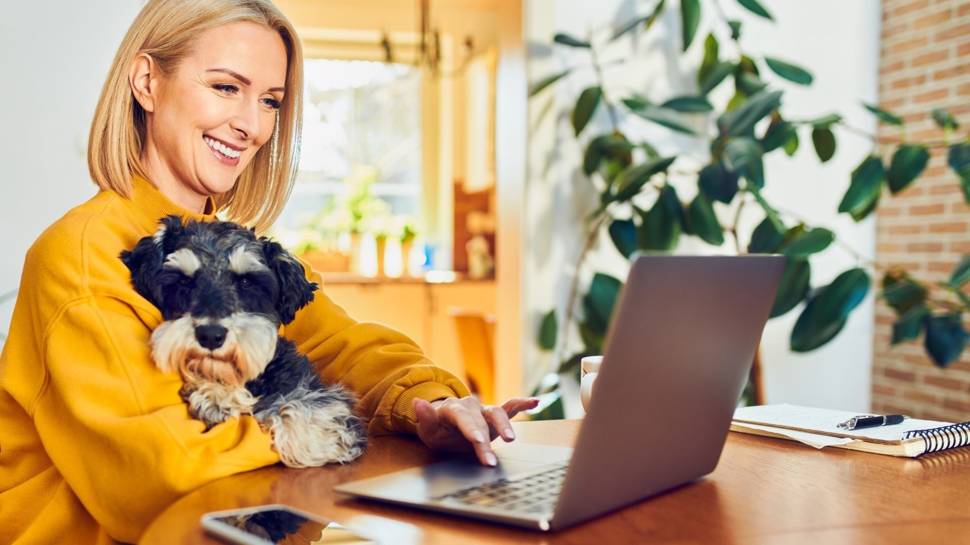 A smiling lady holding a dog works on a laptop computer.
