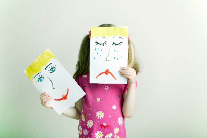 A child holds two pictures of happy and sad faces. She is holding the sad face in front of her face.